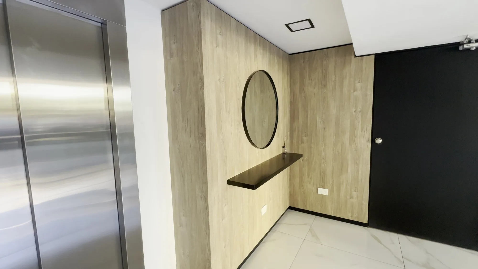 Hall de acceso revestido en madera, con espejo redondo y ascensor de acero inoxidable